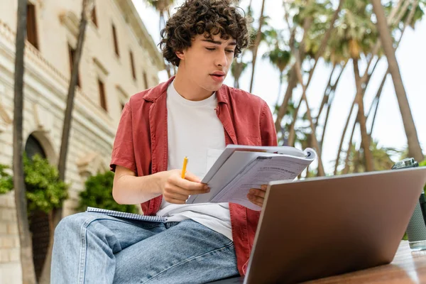 Portrait of trendy dressed male student looking at the notebook and laptop attentively while learning at urban bench in city