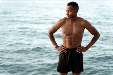 Afrika kökenli, sportif vücutlu ve iyi görünümlü bir adam. Arka planda dalgalı bir denizle kıyıda durup düşünürken başka yöne bakıyor ve düşünüyor.