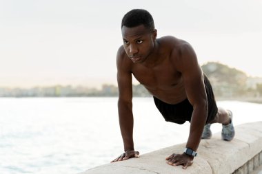 Kendine güvenen Afro-Amerikan tişörtsüz adam plajda konsantre yüzüyle şınav çekiyor. Spor ve eğlence konsepti