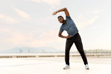 Spor kıyafetleri içinde konsantre bir erkek. Güneşli bir günde denize yakın kumsalda dikilip gözlerini kaçırarak esneme egzersizleri yapıyor. 