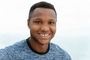 Afro-Amerikan bir adamın portre görüntüsü sportif vücutlu ve iyi durumda kameraya bakıyor ve arka planda deniz kenarında dururken poz veriyor.