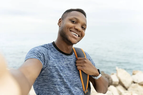 Akıllı telefonunda sırt çantasıyla selfie çekerken kameraya gülümseyen neşeli melez ırk adamı. İnsan duyguları kavramı