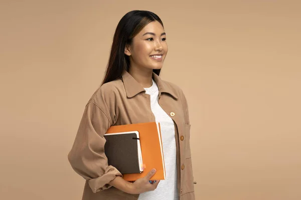 Pretty asian woman student holding books or paper organizers, looking away with smile. Indoor studio shot isolated on beige background