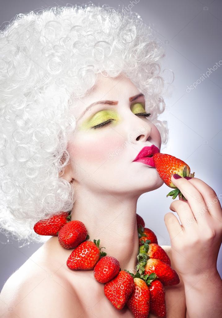 Creative makeup beauty shot of model with strawberries, artistic edit .Woman with strawberry necklace, wig and makeup professionally posing in studio.Beauty with strawberry