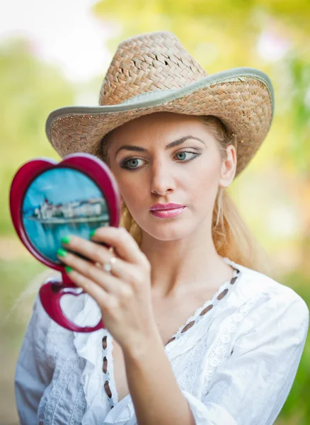 Bella donna con cappello di paglia e specchio. Giovane donna guardando nello specchio cosmetico. Bella bella donna che fa il trucco davanti allo specchio . — Foto Stock