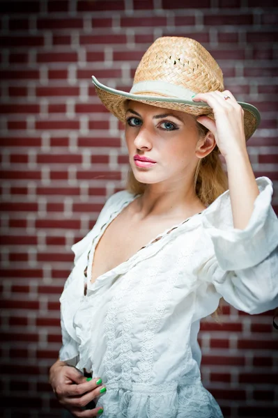 Atractiva chica rubia con sombrero de paja y blusa blanca. Hermosa mujer joven con sombrero de paja y kaftan blanco contra una pared de ladrillo. Hermosos ojos azules rubios mientras usa un sombrero de vaquero de paja. . —  Fotos de Stock