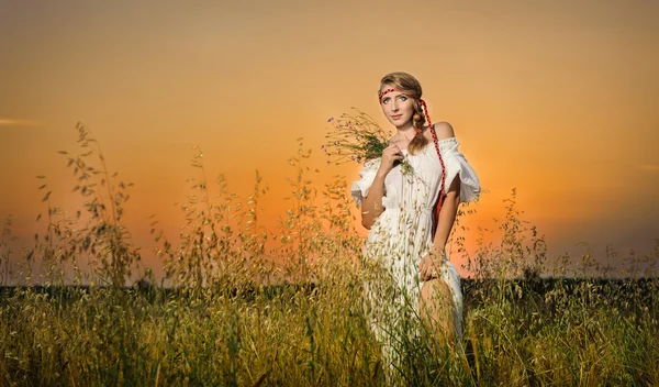 Giovane donna in piedi su un campo di grano con alba sullo sfondo.Ritratto di ragazza in campo.Romantica giovane donna in posa all'aperto.Donna attraente in abito bianco in campo di grano giallo all'alba . — Foto Stock