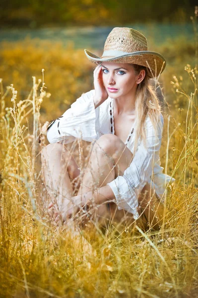 Modeporträt Frau mit Hut und weißem Hemd sitzt auf einem Heustapel.Sehr nette blonde Frau sitzt draußen auf dem gelben Gras mit Hut — Stockfoto