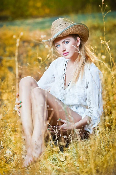 Modeporträt Frau mit Hut und weißem Hemd sitzt auf einem Heustapel.Sehr nette blonde Frau sitzt draußen auf dem gelben Gras mit Hut — Stockfoto