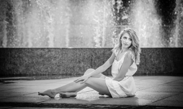 Jolie fille en robe courte blanche assise sur le parapet près de la fontaine dans la journée la plus chaude d'été.Fille avec robe partiellement humide jouant avec l'eau.Portrait de belles femmes blondes près de la fontaine — Photo