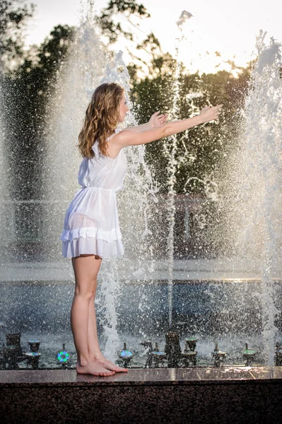 Приваблива дівчина в білій короткій сукні, що сидить на парапеті біля фонтану в літній найспекотніший день. Дівчина з сукнею, частково мокра, грає з водою. Портрет красивих блондинок біля фонтану — стокове фото
