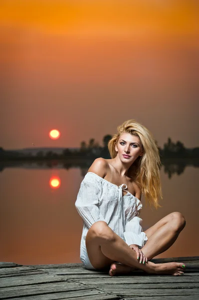 Bella ragazza con una camicia bianca sul molo al tramonto.Donna sexy con lunghe gambe sedute su un molo. Immagine a colori di una ragazza di bellezza seduta su un molo, affacciata su un lago — Foto Stock