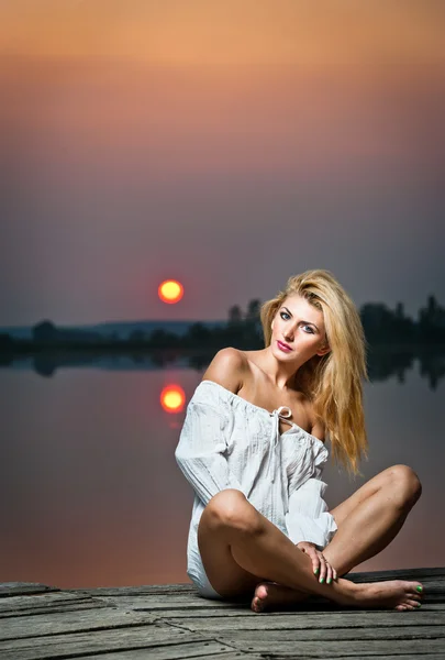 Menina bonita com uma camisa branca no cais ao pôr do sol.Sexy mulher com pernas longas sentadas em um cais. Imagem colorida de uma menina de beleza sentada em um cais, com vista para um lago — Fotografia de Stock