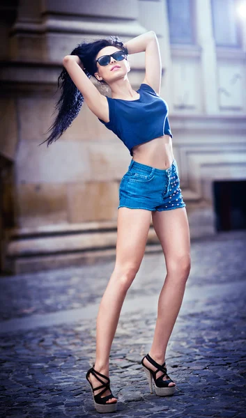 Porträtt av en vacker sexig kvinna med jeansshorts och solglasögon i urban bakgrund. attraktiv kvinnlig brunett kvinna poserar i blå jeans kort i stadsbilden .young kvinna i blå jeans kort — Stockfoto