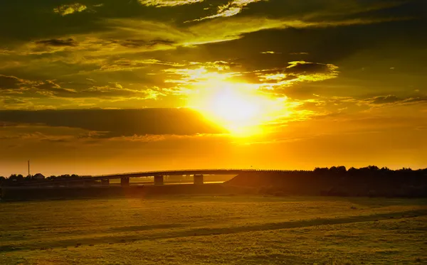 Puesta de sol con cielo dramático sobre campo verde agrícola con un puente. Campo verde y puente al atardecer. Campo de trigo al atardecer —  Fotos de Stock