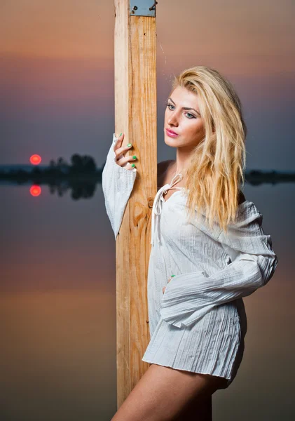 Menina bonita com uma camisa branca no cais ao pôr do sol.Sexy mulher com pernas longas sentadas em um cais. Imagem colorida de uma menina de beleza sentada em um cais, com vista para um lago — Fotografia de Stock