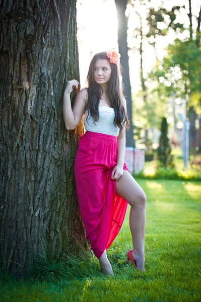 Longitud completa de la joven mujer caucásica con falda larga roja de pie cerca del árbol al aire libre.Retrato romántico de la mujer en el bosque —  Fotos de Stock