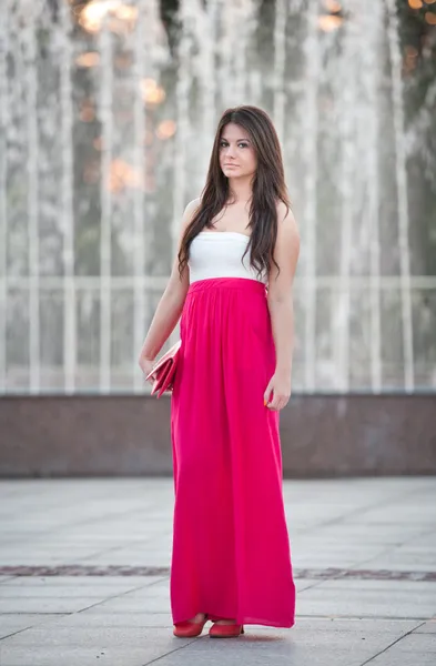 Full length of young caucasian female with long red skirt standing in front of a fountain in a summer day — Stock Photo, Image