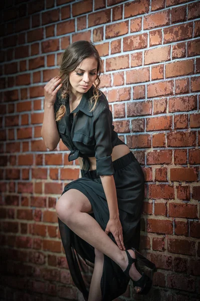 Очаровательная молодая брюнетка в черном платье и на высоких каблуках возле стенки брика. — стоковое фото