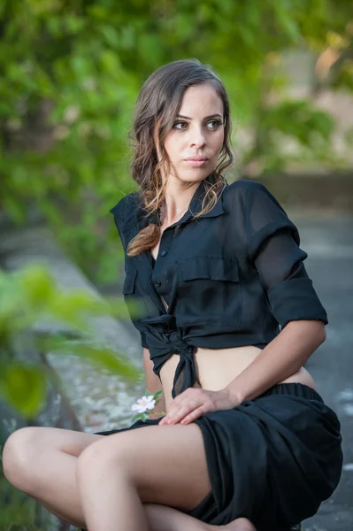 Encantadora mujer morena joven en vestido negro y tacones altos sentado en la pared de ladrillo. hermosa mujer joven con piernas largas relaxing.Full longitud retrato de una mujer con el pelo largo y camisa negra —  Fotos de Stock