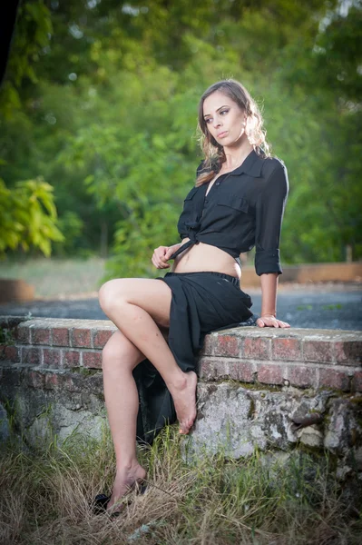 Charming young brunette woman in black dress and high heels sitting on brick wall.Sexy gorgeous young woman with long legs relaxing.Full length portrait of a woman with long hair and black shirt — Stock Photo, Image