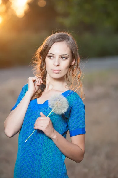Portrait of a brunette young woman in blue dress with a big dandelion on a background of warm sunset. summer, outdoors.Beautiful girl with dandelion enjoying the summer sun outdoors in the park — Stock Photo, Image