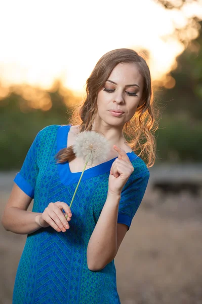 Ritratto di giovane donna bruna in abito blu con un grande dente di leone su uno sfondo di caldo tramonto. estate, all'aperto.Bella ragazza con dente di leone godersi il sole estivo all'aperto nel parco — Foto Stock