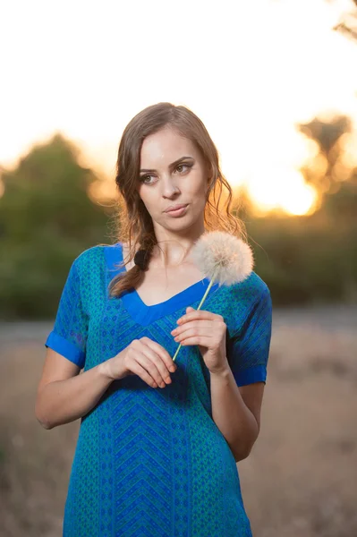 Portrait d'une jeune femme brune en robe bleue avec un gros pissenlit sur fond de coucher de soleil chaud. été, extérieur.Belle fille avec pissenlit profiter du soleil d'été en plein air dans le parc — Photo