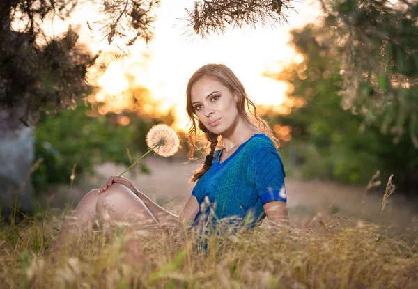Retrato de uma jovem morena em vestido azul com um grande dente de leão em um fundo de pôr do sol quente. verão, ao ar livre.Menina bonita com dente de leão apreciando o sol de verão ao ar livre no parque — Fotografia de Stock