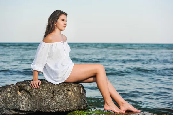 Kaukasisches Teenie-Mädchen im Bikini und weißem Hemd auf Lavafelsen am Meer liegend. Teenager-Mädchen in weißem Hemd auf einem Felsen im Meer sitzend.. — Stockfoto