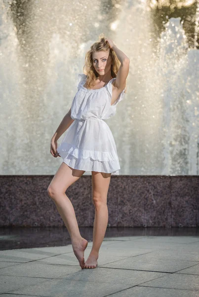 Attractive girl in white short dress sitting in front of a fountain in the summer hottest day. Girl with dress partly wet dancing. Beautiful blonde women near the fountain in a ballet position — Stock Photo, Image
