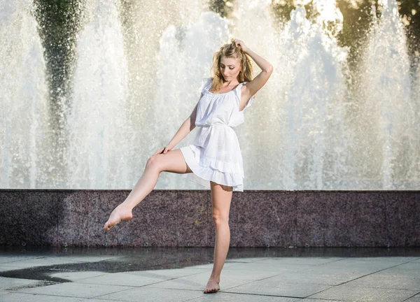 Attractive girl in white short dress sitting in front of a fountain in the summer hottest day. Girl with dress partly wet dancing. Beautiful blonde women near the fountain in a ballet position — Stock Photo, Image