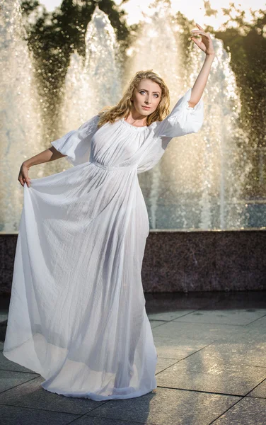 Attractive girl in white long dress sitting in front of a fountain in the summer hottest day. Girl with dress partly wet posing near a fountain. Blonde women near the fountain in a ballet position — Stock Photo, Image