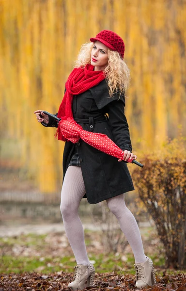 Jovem atraente em uma sessão de moda de outono. Menina bonita e elegante com guarda-chuva vermelho, boné vermelho e cachecol vermelho no parque. Mulheres loiras com acessórios vermelhos posando ao ar livre — Fotografia de Stock