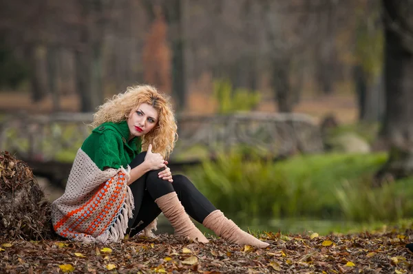 Beautiful woman posing in park during autumn season. Blonde girl wearing green blouse and big shawl posing outdoor. Long fair hair girl with green sweater under a shawl relaxing in autumnal park. — Stock Photo, Image