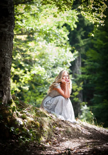 可爱的年轻女士穿着雅致的白色连衣裙，享受光束天体在魔法森林里的她脸上。与白色裙子的漂亮金发仙女。在树林中的迷人公主 — 图库照片
