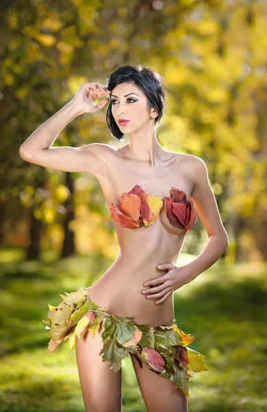 Foto di sensuale ragazza bruna in posa in un bellissimo parco autunnale vestita con un abito fatto da foglie arrugginite. Elegante femmina ricoperta da foglie autunnali. Giovane donna godendo di una giornata di sole autunnale nel parco — Foto Stock