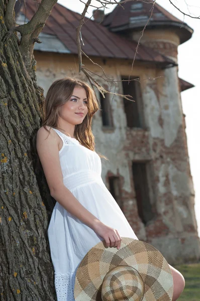 Jolie jeune femme posant devant la ferme. Très jolie fille blonde avec robe courte blanche tenant un chapeau. Romantique jeune femme posant en plein air dans le domaine — Photo