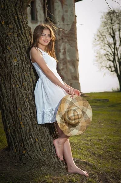 Una mujer joven y bonita posando frente a la granja. Chica rubia muy atractiva con vestido corto blanco sosteniendo un sombrero. Romántica joven posando al aire libre en el campo —  Fotos de Stock