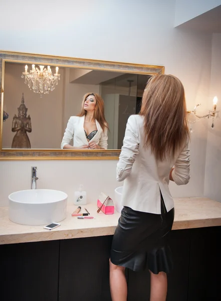 Sensual elegant woman in office outfit looking into a large mirror. Beautiful and sexy blonde young woman wearing an elegant white jacket and a black midi skirt posing in a mirror. Fashionable model. — Stock Photo, Image