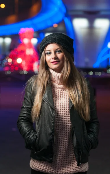 Senhora elegante vestindo boné e casaco preto ao ar livre em paisagens xmas com luzes azuis no fundo. Retrato de jovem mulher bonita com cabelo longo e claro posando sorrindo no estilo de inverno . — Fotografia de Stock