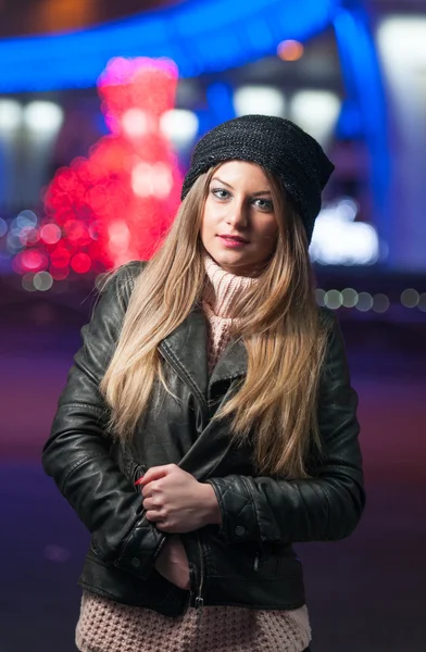 Modische Dame mit Mütze und schwarzer Jacke draußen in der Weihnachtslandschaft mit blauem Licht im Hintergrund. Porträt einer jungen schönen Frau mit langen blonden Haaren, die lächelnd im Winterstil posiert. — Stockfoto