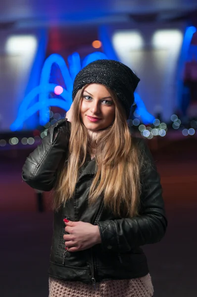 Modieuze dame dragen GLB en zwarte jas buiten in xmas landschap met blauwe lichten op achtergrond. Portret van jonge mooie vrouw met lang eerlijke haar poseren glimlachend in winter stijl. — Stockfoto