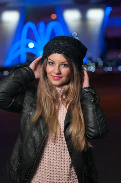 Fashionable lady wearing cap and black jacket outdoor in xmas scenery with blue lights in background. Portrait of young beautiful woman with long fair hair posing smiling in winter style. — Stock Photo, Image