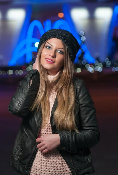 Fashionable lady wearing cap and black jacket outdoor in xmas scenery with blue lights in background. Portrait of young beautiful woman with long fair hair posing smiling in winter style. — Stock Photo, Image