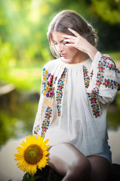 Giovane ragazza che indossa camicetta tradizionale rumena che tiene un colpo di girasole all'aperto. Ritratto di bella ragazza bionda con fiore giallo brillante. Bella donna guardando un concetto di armonia floreale — Foto Stock