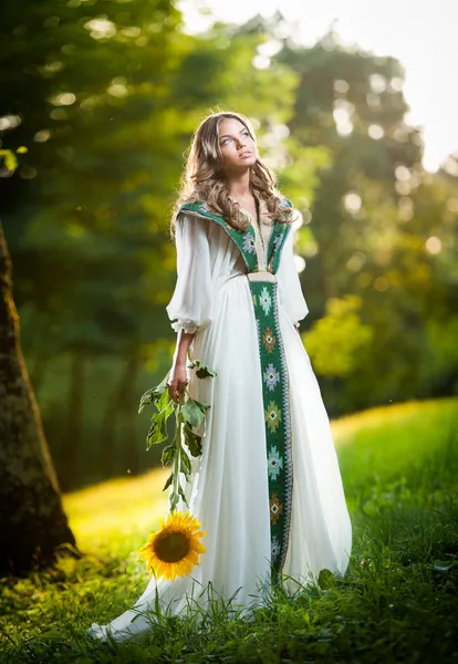 Jovem mulher vestindo um longo vestido branco segurando um girassol tiro ao ar livre. Retrato de menina loira bonita com flor amarela brilhante na floresta. Menina atraente com cabelo longo - cenário de conto de fadas — Fotografia de Stock