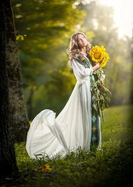 Mujer joven con un vestido blanco largo sosteniendo girasoles tiro al aire libre. Retrato de hermosa chica rubia con flores de color amarillo brillante ramo. Chica atractiva con el pelo largo - paisaje de cuento de hadas —  Fotos de Stock