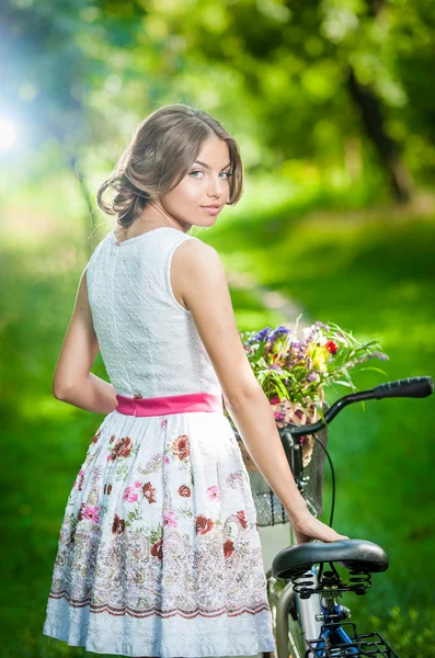 Belle fille portant une jolie robe blanche s'amusant dans le parc avec vélo. Style de vie sain en plein air concept. Paysages anciens. Jolie fille blonde avec look rétro avec vélo et panier avec des fleurs — Photo