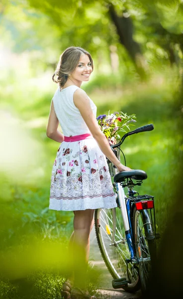 Hermosa chica con un bonito vestido blanco que se divierte en el parque con la bicicleta. Concepto de estilo de vida saludable al aire libre. Paisaje vintage. Linda chica rubia con look retro con bicicleta y cesta con flores — Foto de Stock